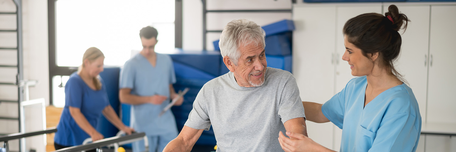 Cheerful senior man using parallel bars to walk and physiotherapist smiling at him